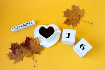 Calendar for September 16 : the name of the month in English, cubes with the number 16,a white heart-shaped cup on a saucer with tea, maple leaves, yellow background, top view