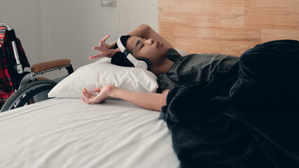 Asian handicapped teenager boy listening music video in the morning on bed and wheelchair in room, Lifestyle activities of disabled child using technology and remote learning.