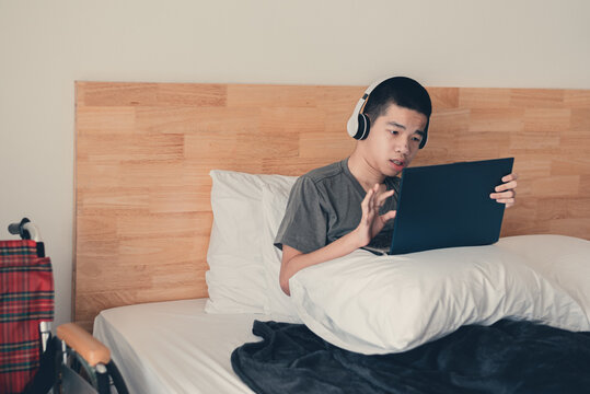 Asian handicapped teenager boy using computer and listening music video in the morning on bed and wheelchair in room, Lifestyle activities of disabled child using technology and remote learning.