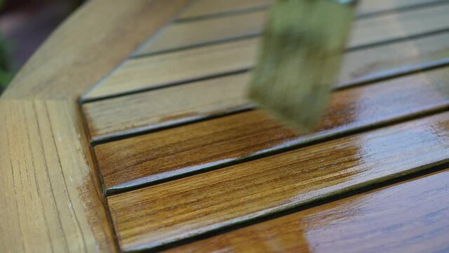 Extreme closeup slow motion of paintbrush applying varnish to teak table furniture outside.