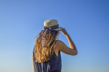 Blue sky and a girl with a dress and a hat standing backwards - obrazy, fototapety, plakaty