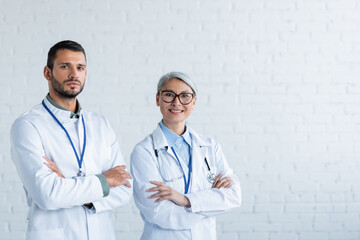 asian doctor smiling near serious colleague while standing with crossed arms