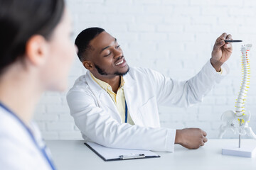smiling african american doctor pointing with pen at spine model near blurred colleague