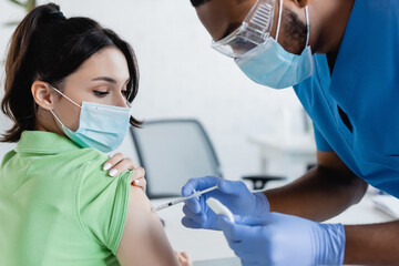 african american physician in medical mask and goggles vaccinating young woman in clinic