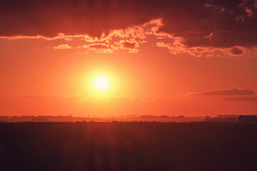 Hot red sun behind the silhouettes of city buildings. Summer sunset in the sky haze with night houses