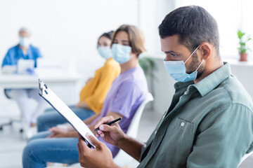 young man in medical mask writing on clipboard near blurred people and nurse, vaccination concept