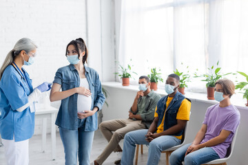 mature asian nurse vaccinating young pregnant woman near multiethnic patients sitting in medical masks