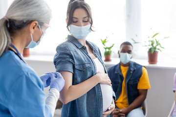 mature nurse vaccinating pregnant woman near blurred african american man in medical mask