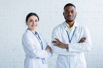 positive multiethnic doctors in white coats standing with crossed arms and smiling at camera