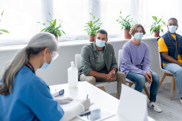 blurred nurse writing near multiethnic people in medical masks waiting in vaccination center