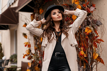Fashionable beautiful woman model in elegant trendy clothes with hat and gray coat walking in the city with yellow autumn foliage
