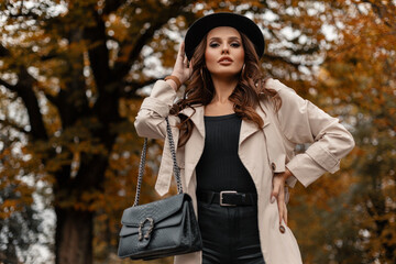 Beautiful young elegant girl with curly hair in fashionable clothes with a stylish hat and a leather black handbag walks on the street against the background of trees with autumn foliage