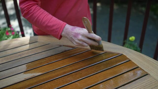 Closeup of paintbrush applying varnish to teak table furniture outside.
