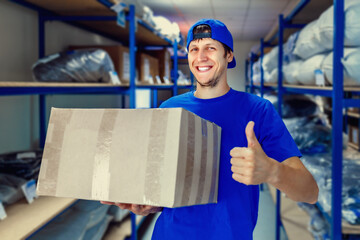 A young positive warehouse worker holds a cardboard box in his hands and smiles. The concept of...