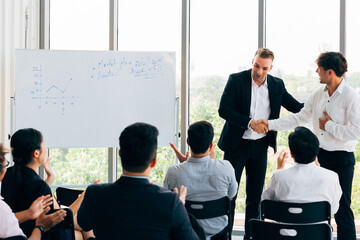 Caucasian business manager introduces new Asian businessman in front of seminar meeting with many employees listening in company office setting