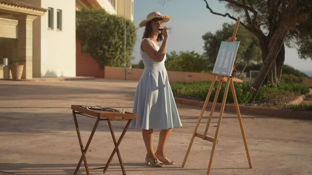 Confident creative Caucasian woman in elegant dress and straw hat drinking red wine admiring picture on easel in sunshine. Wide shot of slim graceful artist standing in sunlight with artwork on canvas
