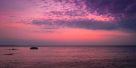 Dramatic overcast clouds, pink sunrays, and reflective ocean surface with a zen-like rock at dawn on Cape Cod in the summer