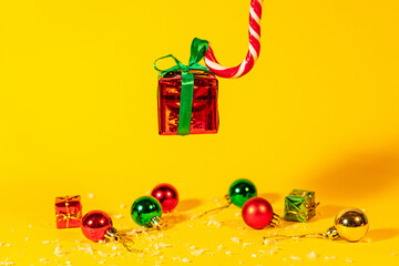 Candy cane lollipop holding a gift box with a Christmas present on a yellow background, Christmas sweets with new year decorations