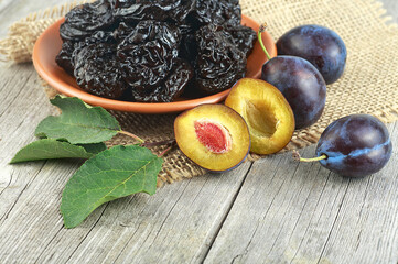Prunes in a clay plate on a wooden table and fresh plums with green leaves. Dried plums, healthy food.