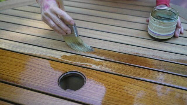 Closeup of paintbrush applying varnish to teak table furniture outside.