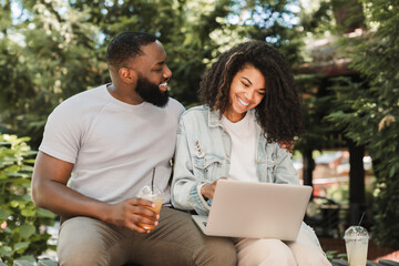 Cheerful african-american young romantic couple friends watching movie comedy online together, surfing Internet, social media, watching webinar online outdoors in city park