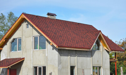 A house under construction with a red asphalt shingled roof, a chimney, attic windows, incomplete...