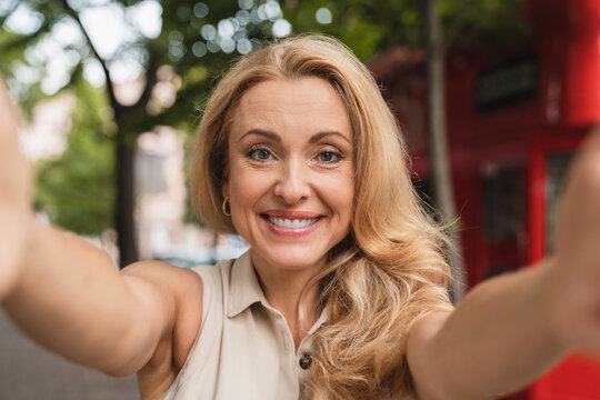 Close up portrait of a blond caucasian middle-aged mature woman businesswoman mother taking selfie on webcam photo having video call online using smart phone outdoors in city park