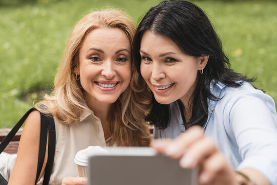 Caucasian mature middle-aged women sisters girlfriends best friends taking selfie photo on smart phone cellphone having video call together while walking in city park outdoors.