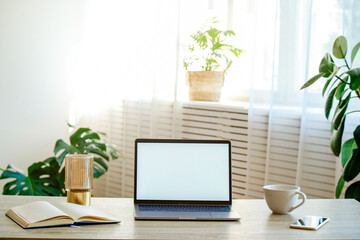 Blank screen laptop, cell phone and a cup on wooden table. Cozy lofty office with decorative palm plants full of natural sunlight. Close up, copy space, background.