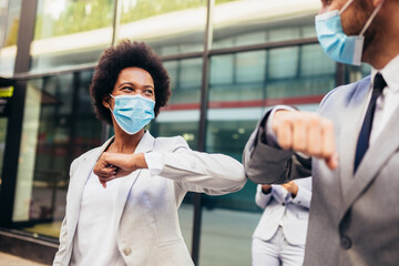 Business man and woman with safety masks greeting with elbow bump in front of office building.