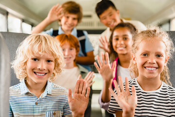 Happy mixed-race multiethnic schoolchildren classmates kids pupils sitting inside school bus waving...