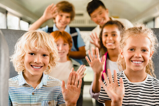 Happy mixed-race multiethnic schoolchildren classmates kids pupils sitting inside school bus waving going back to school lessons classes after summer holidays lockdown. New educational year semester