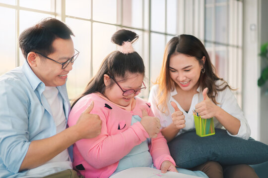 Happy asian family sitting on sofa and  together paint with their daughter down syndrome child in living room at morning time, They show thumbs up , Activity happy family lifestyle concept,