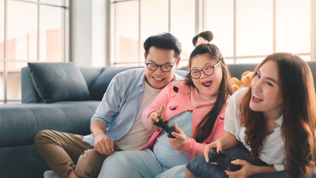 Happy Asian Family Sitting On Floor And Playing Video Games With Their Daughter Down Syndrome Child In Living Room At Morning Time, Activity Happy Family Lifestyle Concept.