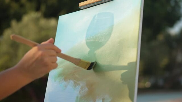 Close-up female hand painting on canvas with silhouette shadow of wine glass reflecting in sunshine. Unrecognizable Caucasian woman enjoying hobby outdoors on sunny summer day. Slow motion