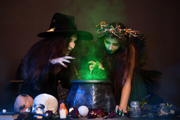 Two witches with disheveled hair brew a magic potion in a black cauldron, standing in a dark room....