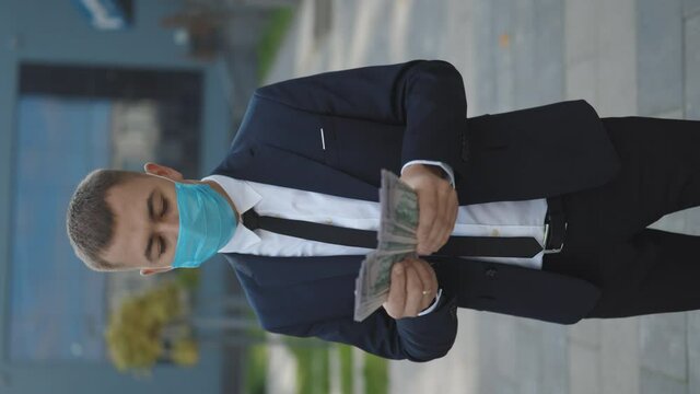 Business Man in Protective Face Mask Counts Cash. Quarantine financial crisis. Young Handsome Rich Man Wearing Stylish Suit Counting Money Standing in the street near Office Building Vertical Video