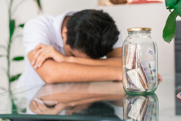 crestfallen young man with his savings in a glass jar on the table, banknotes inside a glass jar. boy worried and bored with his savings. economy and finance concept. Colombian economy - obrazy, fototapety, plakaty