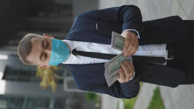 Happy good looking man in business style holding dollars and calculating. Cheerful rich handsome businessman in protective face mask counting money near business center background