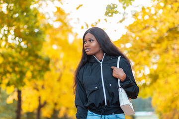 Beautiful young black african woman in a fashionable jacket with a handbag walks in an autumn canada park with bright yellow foliage