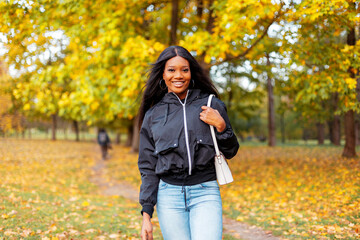 Fresh portrait of a beautiful happy young black girl with a smile in a fashionable casual jacket and jeans with a handbag is walking in an autumn park with golden fall leaves.