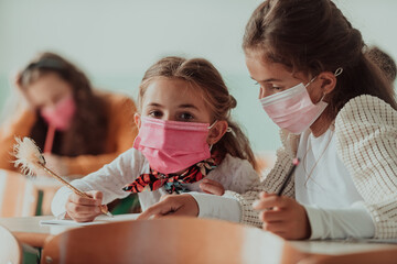 Little girls sitting in school desks while wearing a mask against pandemic corona virus protection. New normal. Education during the Covid-19 pandemic. Selective focus.