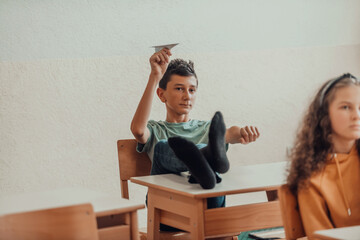 A naughty boy with his feet on a desk sits in the classroom and throws a plane. Selective focus - obrazy, fototapety, plakaty