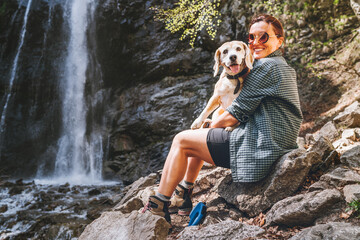 Smiling Female dog owner hugging his friend beagle dog resting near the mountain river waterfall during their together walking in autumn season time. Human and pets or walking in nature concept image.