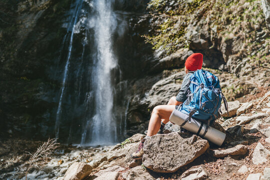 Woman with a backpack in red hat dressed in active trekking clothes sitting near the mountain river waterfall and enjoying the splashing Nature power. Traveling, trekking, and a nature concept image.