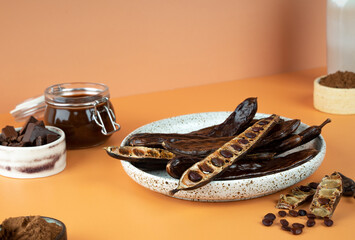 Organic carob pods, seeds in a ceramic plate on a beige background, locust bean healthy food,...
