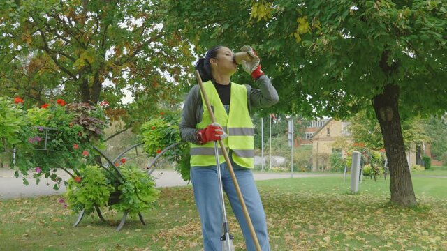 Woman worker drinking coffee and using rake on lawn