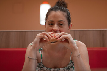 Mujer joven de pelo negro con moño sujetando su hamburguesa de queso y carne sentada en un restaurante vestida de vestido de estampado con una alegria de felicidad
