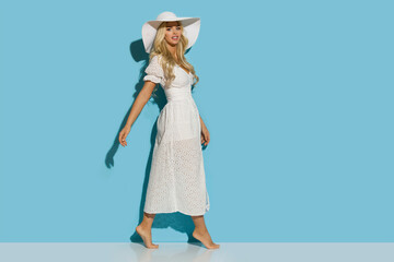 A beautiful woman in a white summer dress and a sun hat is walking barefoot in front of a blue wall