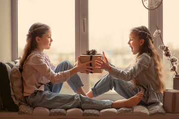 Two beautiful girls are sitting on the windowsill by the window and giving each other a christmas gift